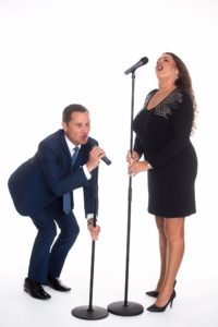 Andy Mack Duo Having Fun With Short And Tall Microphone Stands Against A White Background