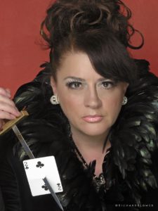 Magic Mandy Stage Magician and Illusionist Dressed in Black Feathers Holding a Sharp Knife With a Playing Card Impaled on it
