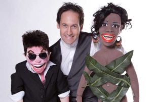 Steve Hewlett Ventriloquist With Sinitta & Simon Cowell Puppets Against A White Background