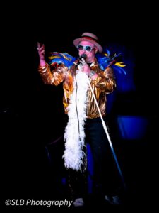 Elton John Tribute Singing Into A Microphone. He Wears A Gold Bomber Jacket And A White Feather Boa. His Shoulders Are Covered In Brightly Coloured Feathers And He Wears a Straw Boater On His Head