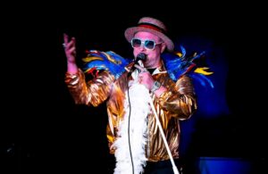 Elton John Tribute Singing Into A Microphone. He Wears A Gold Bomber Jacket And A White Feather Boa. His Shoulders Are Covered In Brightly Coloured Feathers And He Wears a Straw Boater On His Head