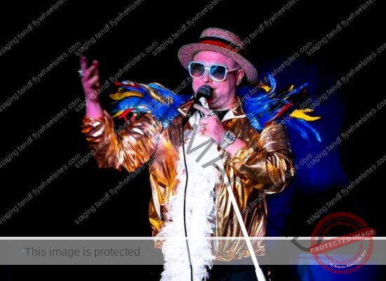 Elton John Tribute Singing Into A Microphone. He Wears A Gold Bomber Jacket And A White Feather Boa. His Shoulders Are Covered In Brightly Coloured Feathers And He Wears a Straw Boater On His Head