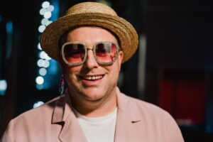 Young Elton John Tribute. He Wears A Pink Jacket , White T Shirt And A Straw Boater. His Glasses Are Studded With Diamantes And He Is Smiling At The Camera.