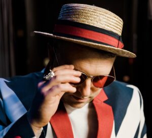 Elton John Tribute Wearing A Union Jack Jacket And Straw Boater. His Glasses Are Orange And There Is A Large Gold Ring On His Finger. His Face Is Slightly Obscured By His Hand.