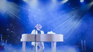 Elton John Tribute Performing Live On A White Baby Grand Piano. He Wears A White Suit And Is Bathed In Numerous White Spotlights From Above. A White Feather Boa Lays On The Lid Of The Piano.