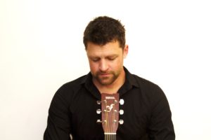 Jay Guitar & Vocals Looking Downwards Against A White Background