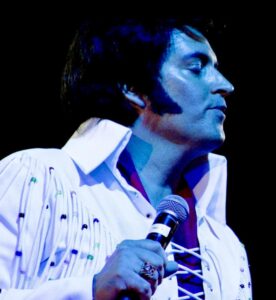 Elvis Presley tribute act performing live. He is dressed in a white and beaded jump suit and is bathed in a blue stage light.