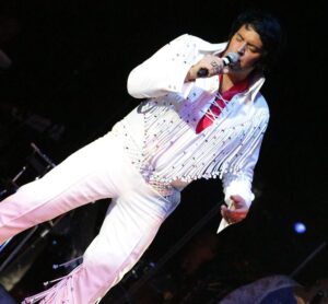 A full length photograph of an Elvis Presley tribute act performing live. He is wearing a white jump suit in the style of Elvis Presley. The photograph is taken at a 45 degree angle, to add to the dynamism of the performance.