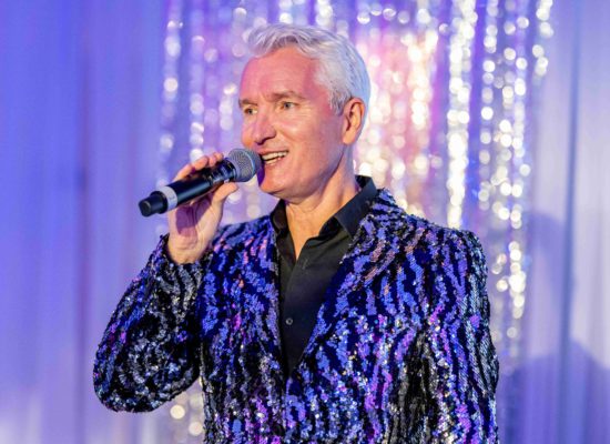 Professional Singer Peter Stanford is singing at a Corporate Event. He is wearing a dazzling silver and black sequinned jacket and stands on stage in front of a backdrop of silver lights which are twinkling.