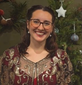 Mitzi Metford is posing in front of a Christmas tree. She wears a vintage gown which is highly decorated with festive sparkle around the neck line. She is smiling directly at the camera.